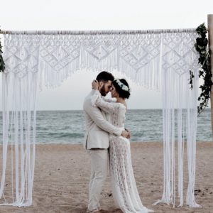 couple de mariés sur la plage, arche, décoration macramé, robe de mariée transparente, costume marié en lin