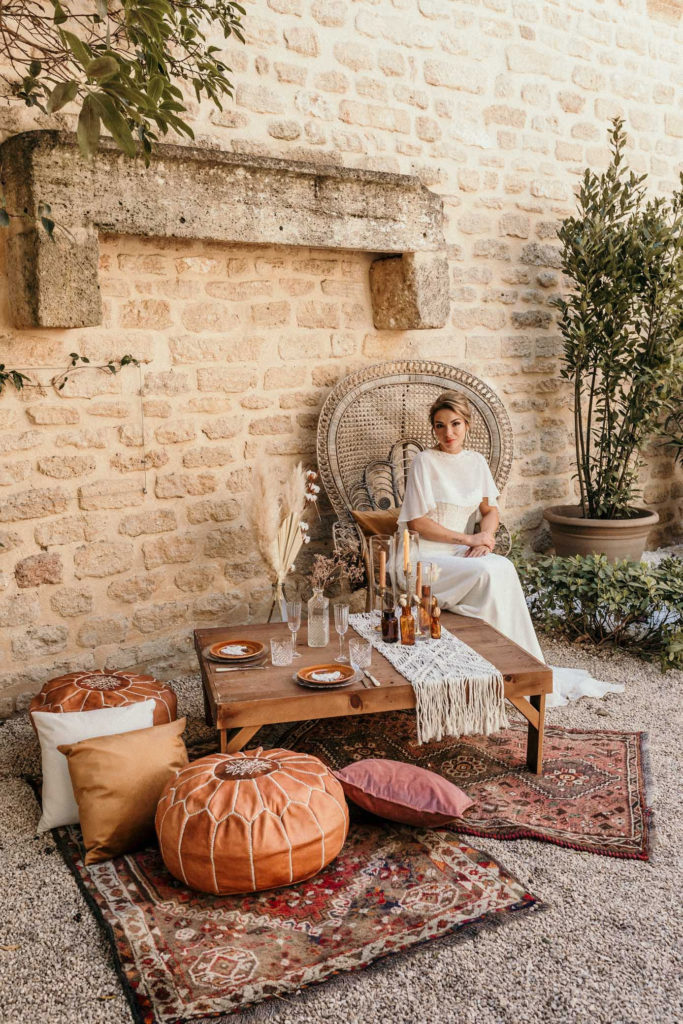 robe de mariée bustier, robe de mariée, fauteuil Emmanuelle rotin, pouf orange cuir, tapis bohème, coussins oranges, mariée assise, chignon mariée, cape robe de mariée, Hôtel Vieux Castillon, inspiration mariage bohème